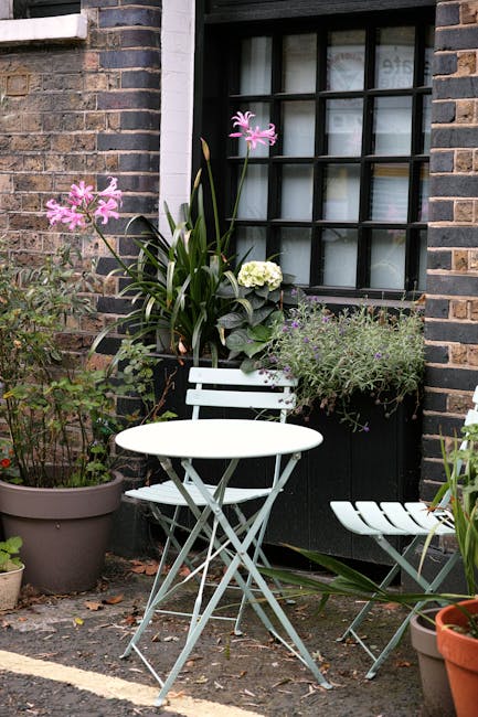 A cozy urban garden scene featuring a small paved patio area with a white metal bistro table and matching folding chair. The terrace is bordered by a brick wall with a large black-framed window, parti