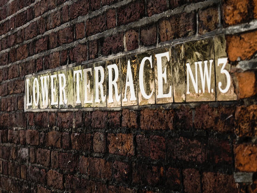 A cozy urban garden scene featuring a small paved patio area with a white metal bistro table and matching folding chair. The terrace is bordered by a brick wall with a large black-framed window, parti